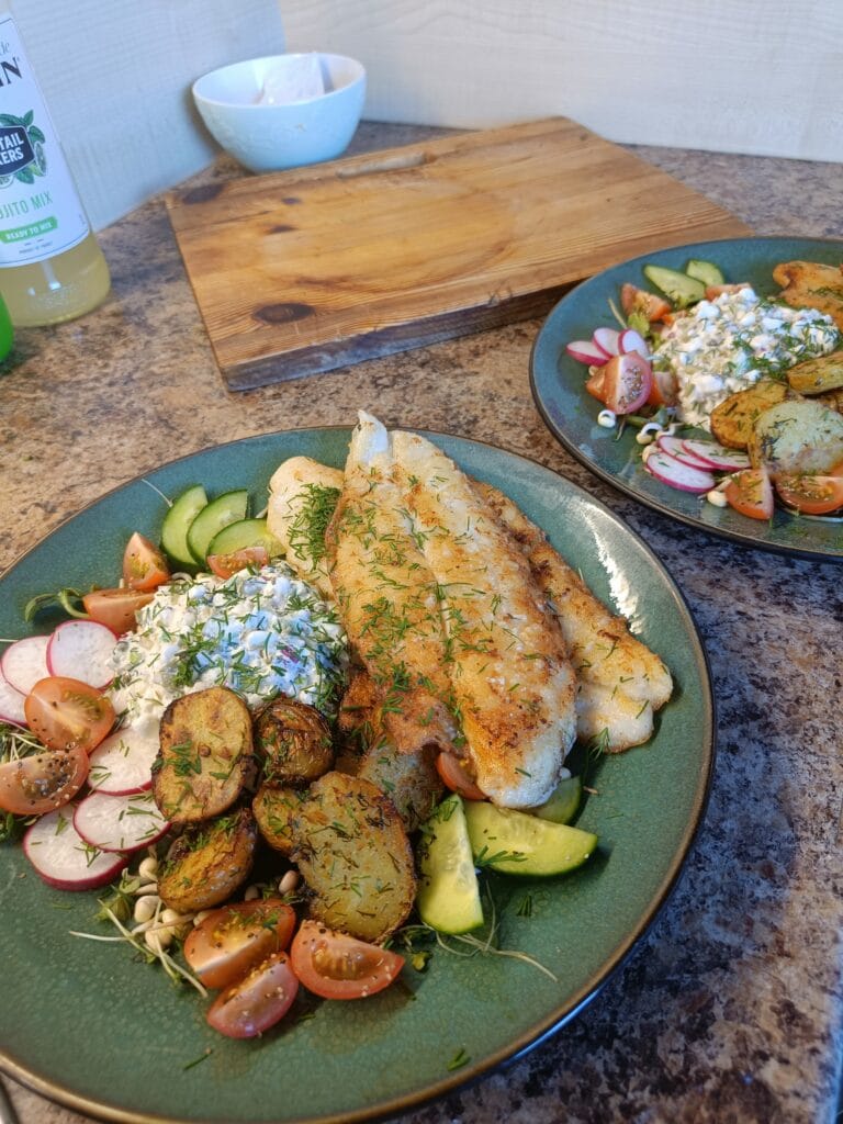 Pan-fried white fish fillets served with roasted potatoes, cucumber, radishes, cherry tomatoes, and a creamy cottage cheese salad on a green plate.