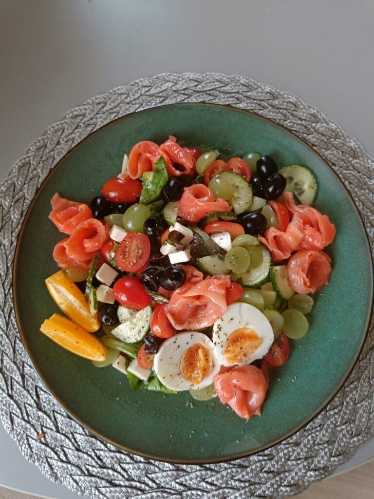 Colourful homemade salad with smoked salmon, soft-boiled eggs, grapes, cherry tomatoes, olives, cucumber, feta cheese, and orange slices served on a green plate.