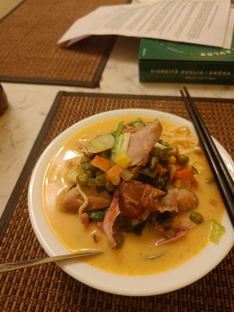 A hearty homemade ramen with vegetables, peas, ham, and creamy broth, served in a bowl with chopsticks and a spoon.