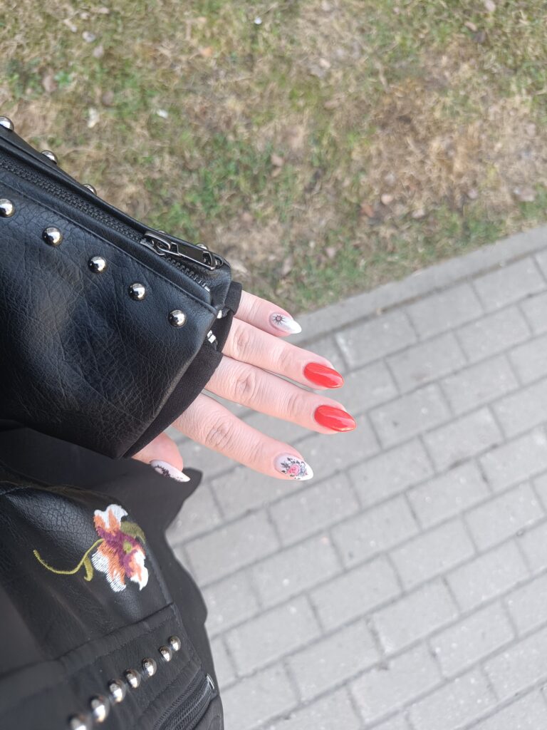 Bright red gel manicure with a floral accent nail, photographed outdoors above a paved walkway.