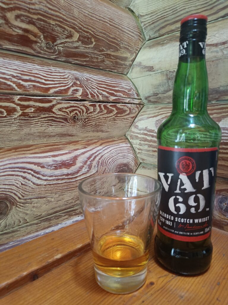 Glass containing amber whisky beside a green VAT 69 whisky bottle on a wooden table against a log cabin wall.