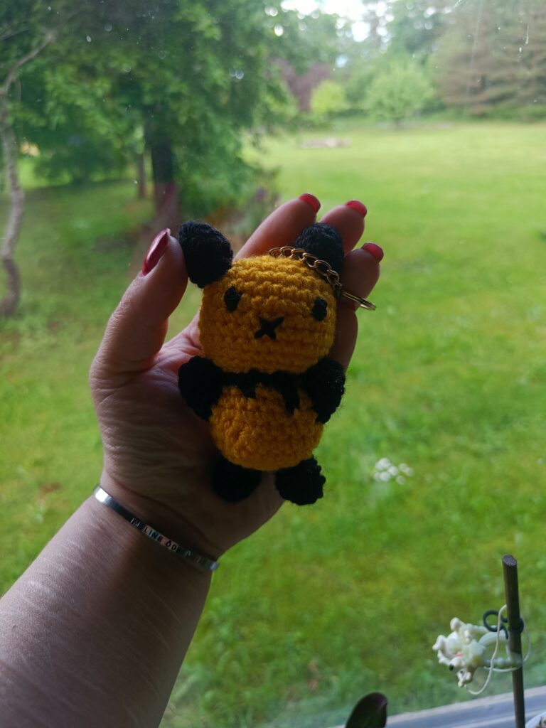 Hand holding a small crocheted bee keychain in front of a green garden.