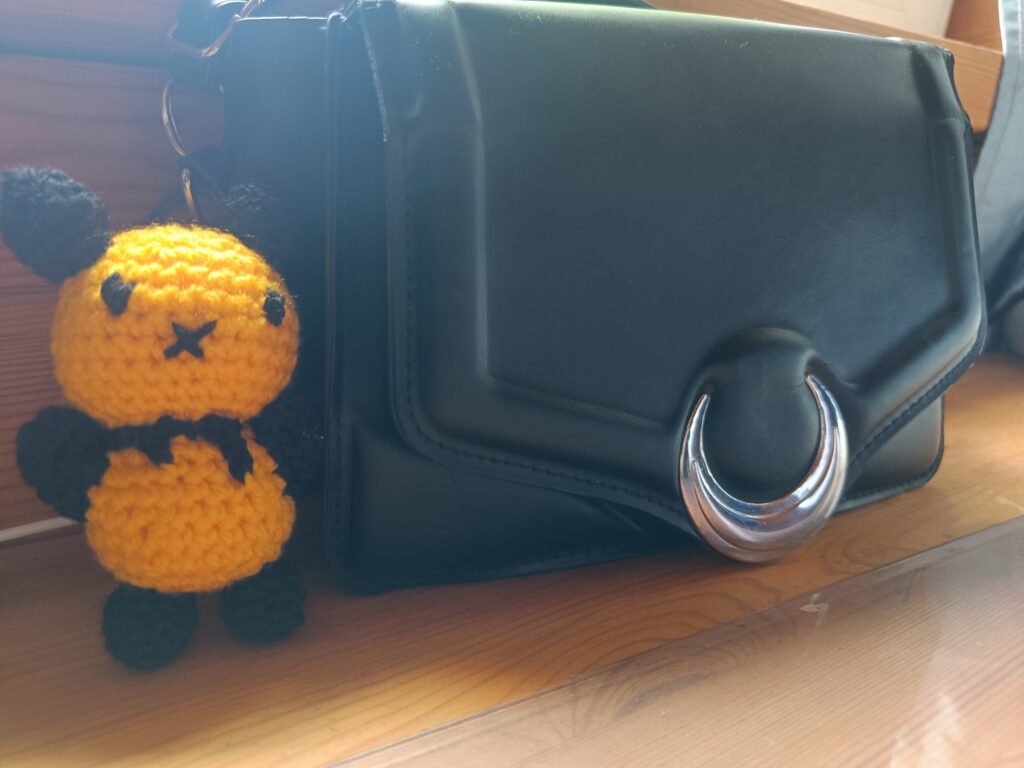 Small crocheted bee toy peeking out from beside a black bag on a wooden floor.