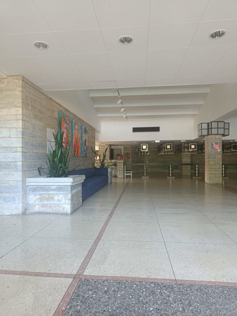 Spacious lobby of the Paide culture house with light stone walls, indoor plants, a blue seating area, and colorful portrait paintings displayed along one wall. The bright, open hall leads toward the entrance and exhibition spaces beyond.