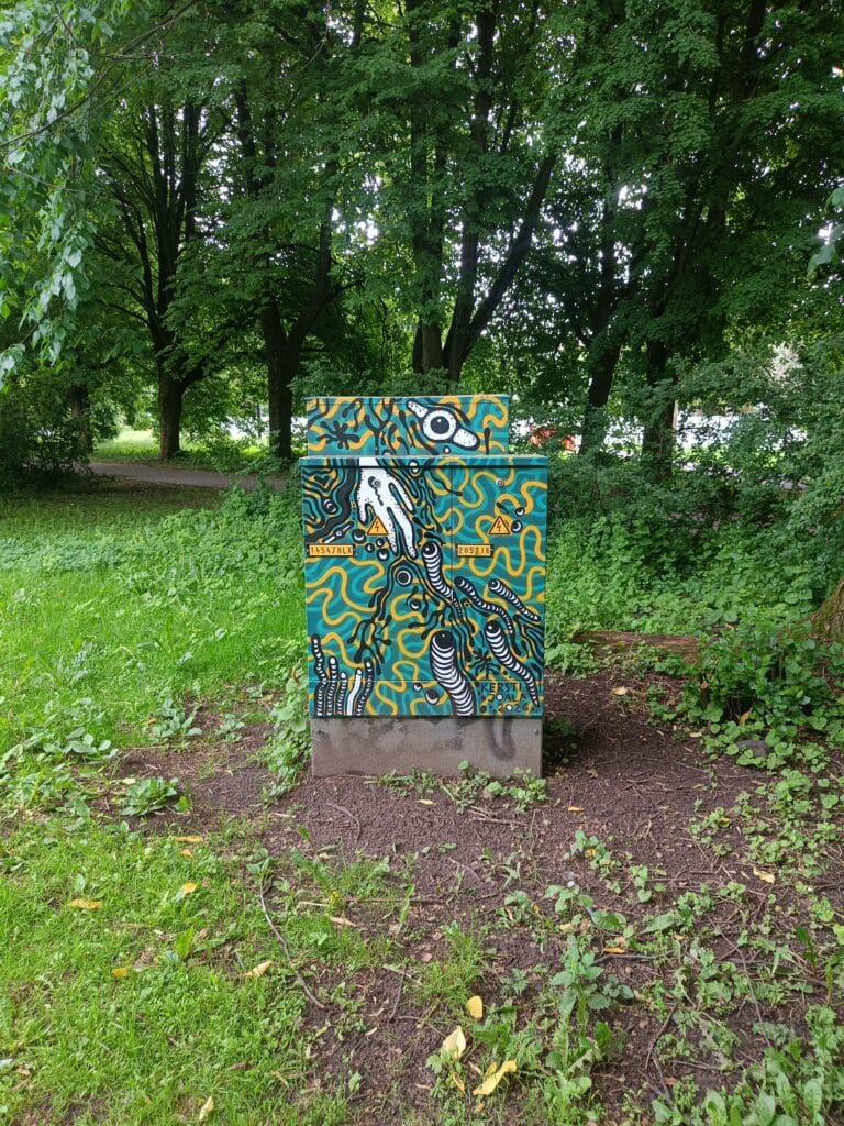 Colourful street art painted on a utility box in a leafy park in Tartu, featuring abstract shapes, tentacle-like forms, and bright turquoise and yellow patterns.