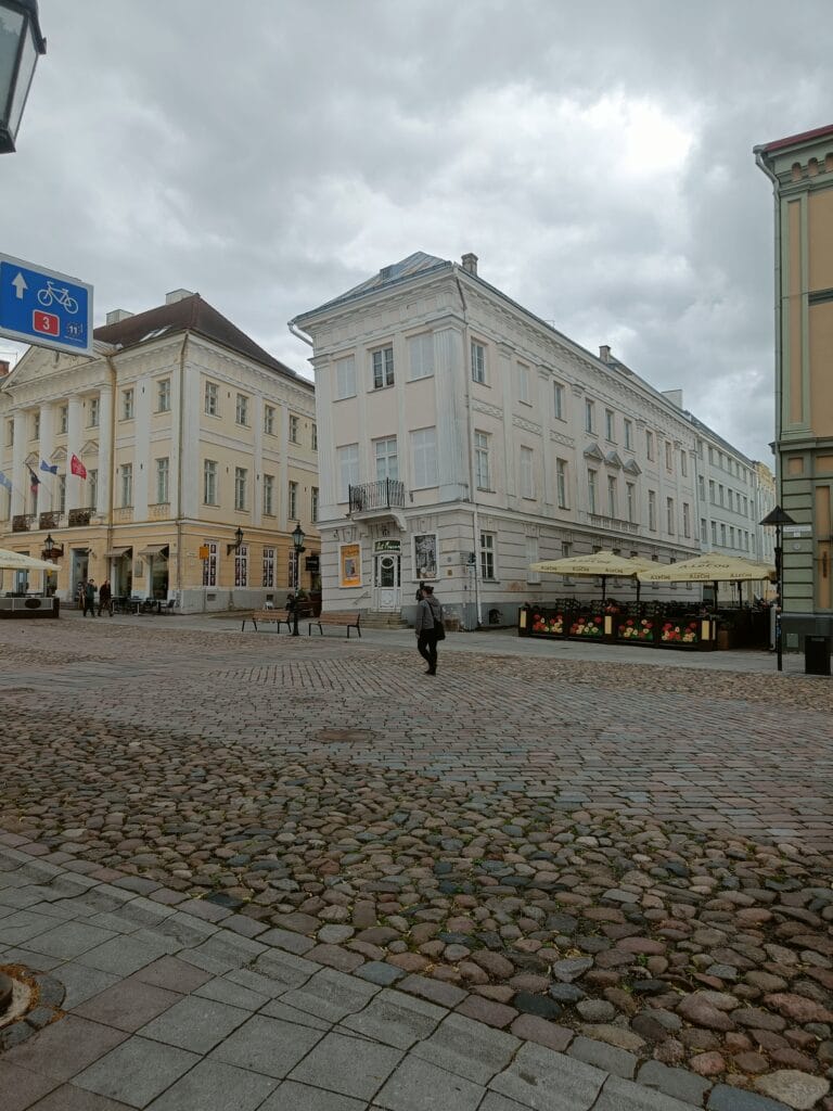 Historic buildings surrounding a cobblestone square in Tartu's city centre, with cafés, pedestrians, and storm clouds gathering overhead. What is cool is that one building looks like the Pisa tower, about to fall over.