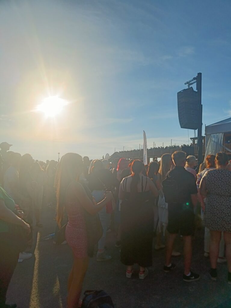 Crowds gathered near the waterfront stage during a bright midsummer evening, with the low sun casting long shadows across the celebration.