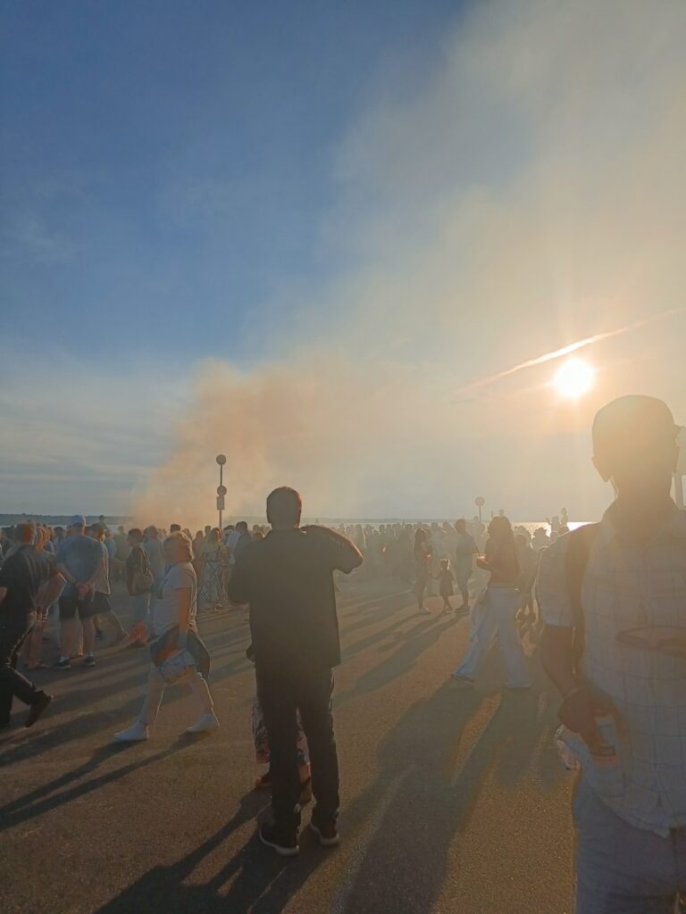 Summer solstice festivities by the sea, with smoke drifting through the warm evening air and crowds bathing in the setting sun.
