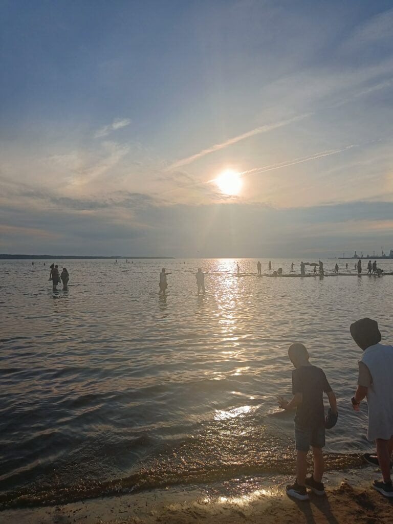 Golden midsummer sunset over the sea, with people cooling off in the shallow water and children playing along the shore.