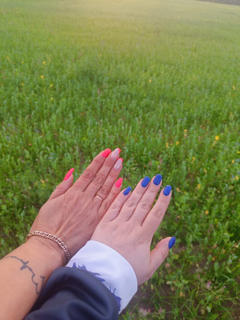 Two hands with brightly coloured gel manicures—one pink and one blue—held out over a green meadow during summer.