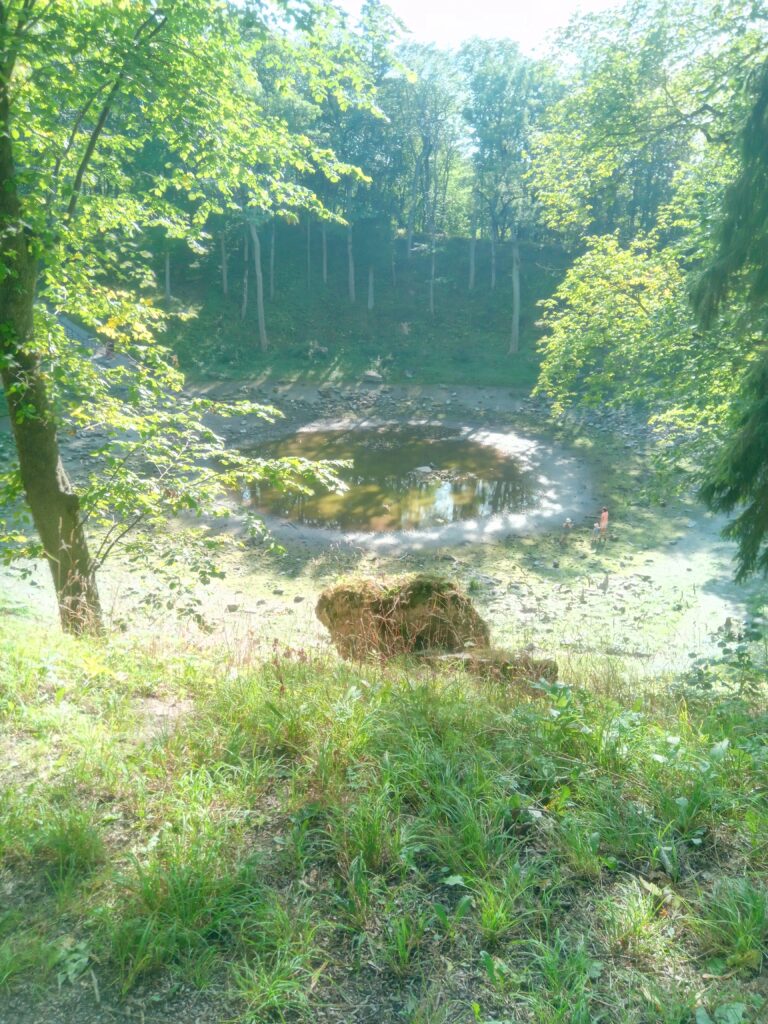 Kaali Crater on Saaremaa, a meteorite impact crater surrounded by trees and grassy slopes, with sunlight reflecting off the water below.