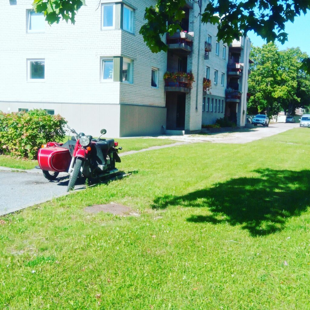 Vintage motorcycle with a red sidecar parked beside an apartment building on a sunny summer day.