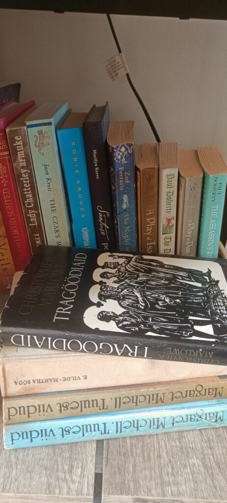 A shelf filled with old and well-loved books, including novels by Agatha Christie, Jaan Kross, Marilyn Kerro, Zoë Ferraris, and Paul Doherty, with several Estonian books stacked underneath.
