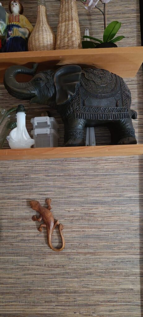 Large decorative elephant figurine on a wall shelf, surrounded by ornaments and a potted orchid, with a small wooden lizard on a wall below.